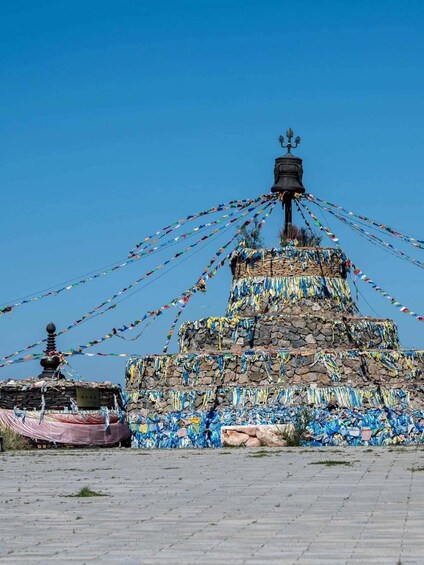 A stone stupa adorned with colorful prayer flags and a bell on top, surrounded by a paved area. 5
