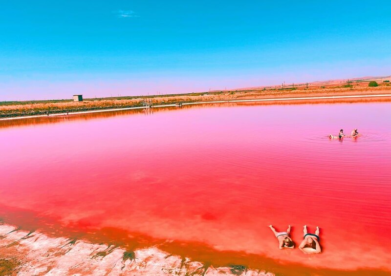 Un extenso cuerpo de agua de color rojizo, un pequeño edificio a lo lejos y tres personas descansando en la orilla. 1