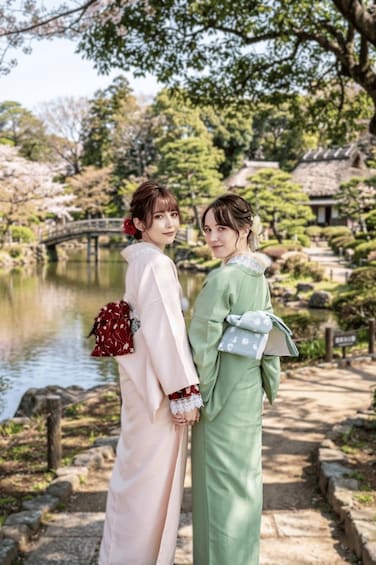Two individuals in traditional attire standing by a pond with a bridge and trees in the background. 2