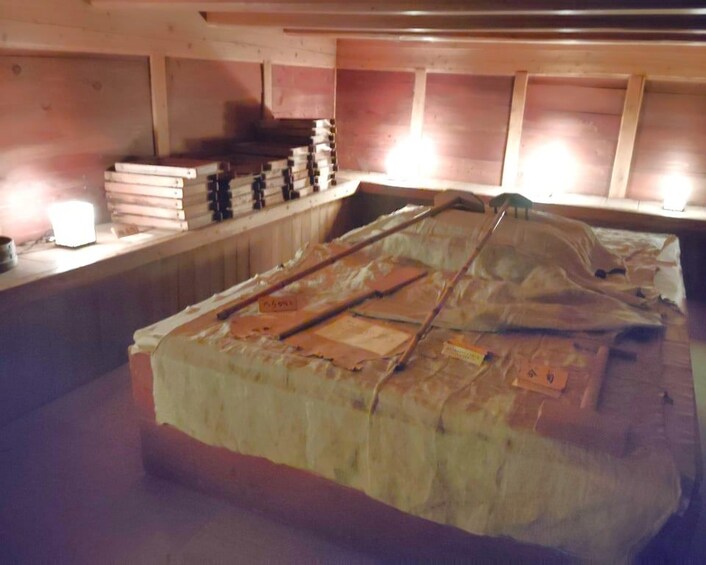A wooden structure with a bed covered in white sheets and blankets, a stack of books on a shelf, and a light source on the left. 2