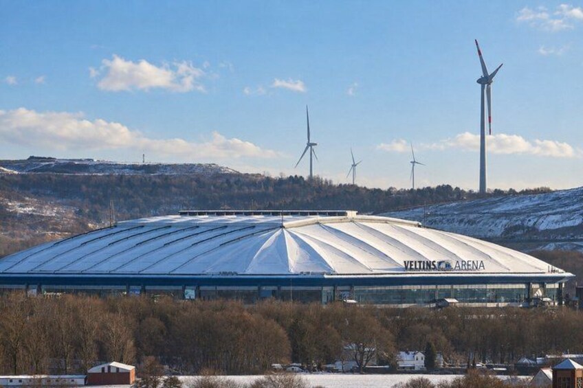 A large stadium named 'Veltins Arena' with a snow-covered roof and wind turbines in the background. 2