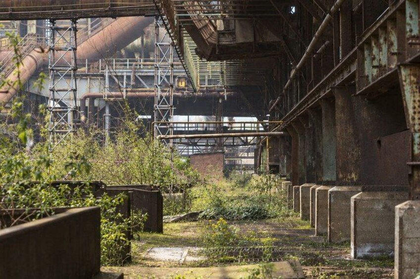 An abandoned industrial area with rusted metal structures, overgrown vegetation, and a dilapidated building. 4