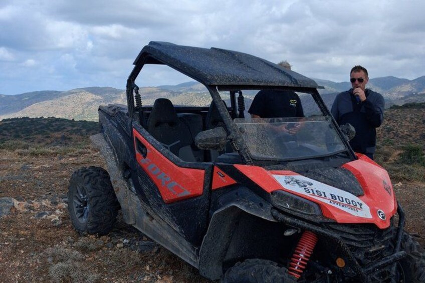 Un vehículo todo terreno rojo y negro con la leyenda 'Sisi Buggy Adventures' en la carrocería, estacionado sobre terreno rocoso con montañas al fondo. 3