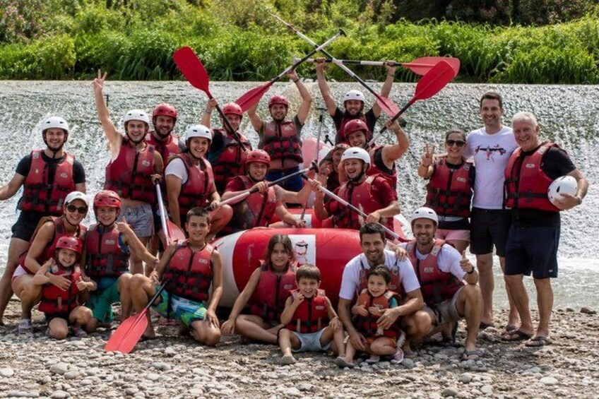 A group of people in red life jackets and helmets, posing with paddles and a red raft on a rocky riverbank. 5