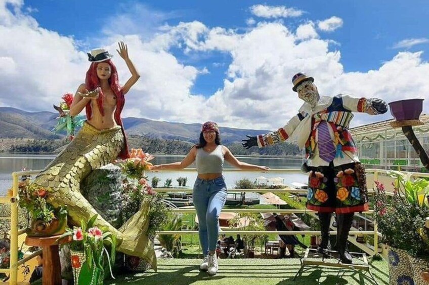 A person in a mermaid costume, another in a colorful traditional outfit, and a third in casual attire, standing on a deck with a scenic lake and mountains in the background. 5