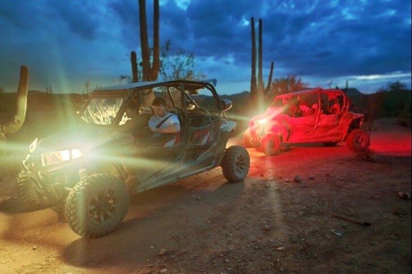 A desert landscape with two off-road vehicles, one with headlights on and another with a red light. 4