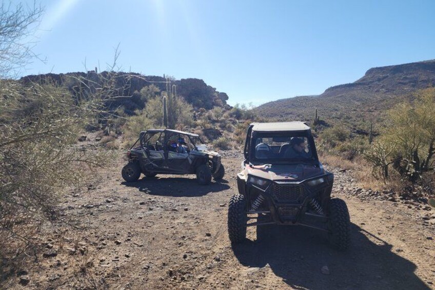 Two off-road vehicles on a dirt path in a desert landscape with sparse vegetation and rocky terrain. 5