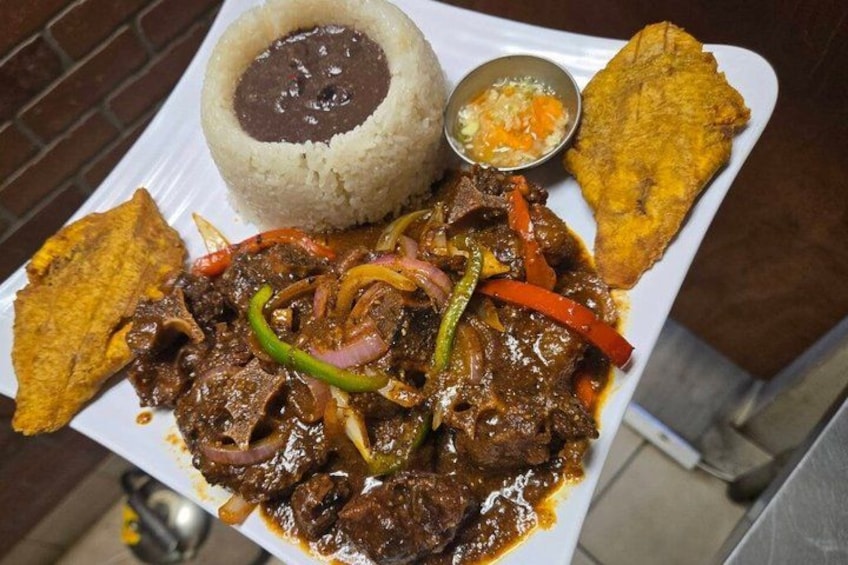 A plate of beef with peppers, a mound of rice, a fried plantain, and a small bowl of sauce. 3