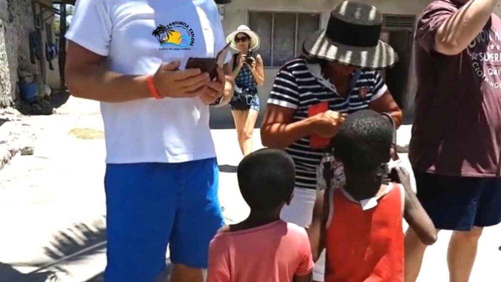 Una persona con camiseta blanca y un logo, de pie en una calle con niños y adultos. 5