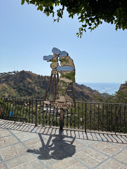 A sculpture of a person with a camera on a tripod, set against a scenic backdrop of hills and a clear sky. 2