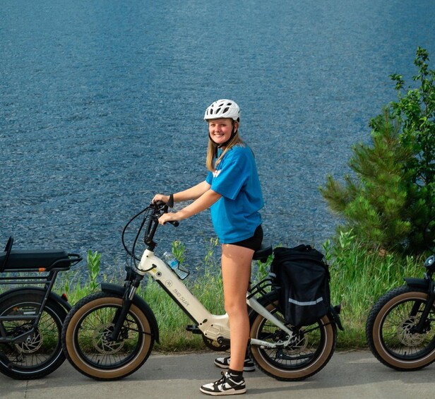A person standing next to a white electric bike with a black bag, near a body of water and greenery. 4