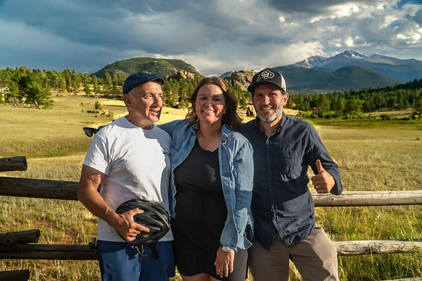 Three people standing by a wooden fence in a grassy field with mountains in the background. 5