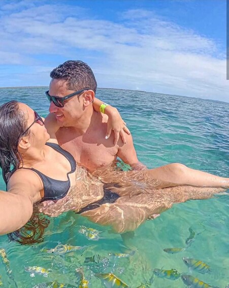Dos personas en un mar azul cristalino, una con traje de baño negro y la otra con una blusa negra, se ve un brazalete en la muñeca. 3