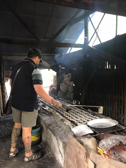 Una persona trabajando con una rejilla metálica, posiblemente para cocinar o crear manualidades, en un entorno rústico al aire libre con un techo improvisado y fuego. 1