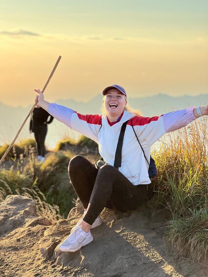 A person sitting on a rocky path with arms outstretched, wearing a cap, a white jacket, and dark pants. 2