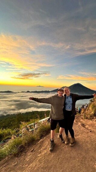 Two people standing on a mountain path with arms outstretched, overlooking a valley with clouds below and a mountain in the distance. 1