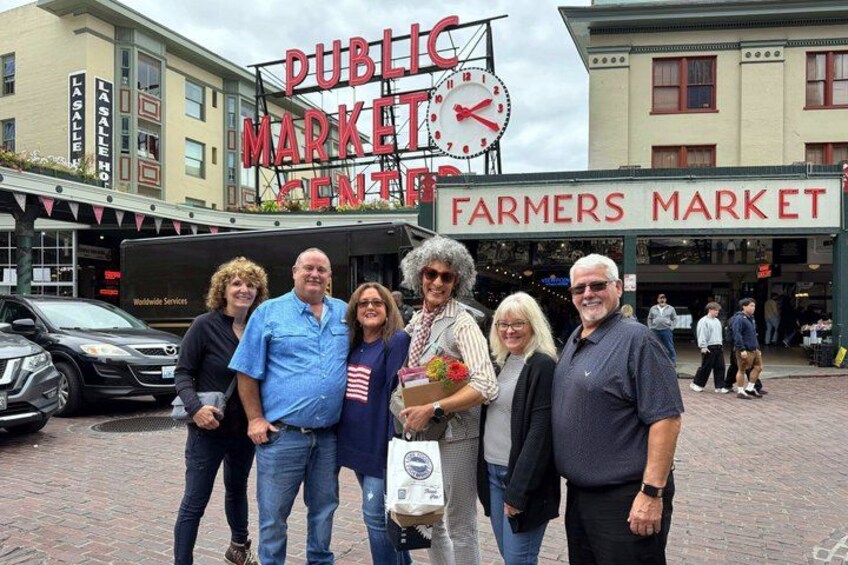 A group of people standing in front of a Farmers Market with a clock and a sign that reads 'Public Market.' 1
