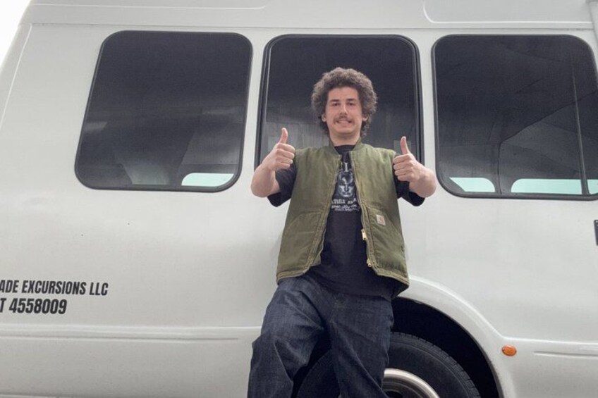 A person sitting on a white vehicle with windows and a logo that reads 'EXCURSIONS LLC' and a phone number. 4