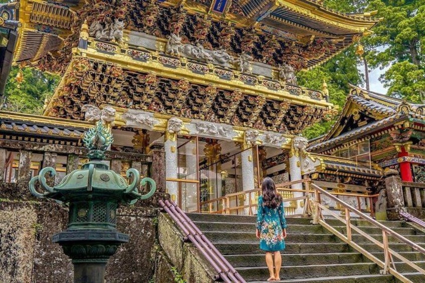 A woman in a blue dress standing on a staircase in front of a traditional ornate building with golden decorations and a green fountain. 1