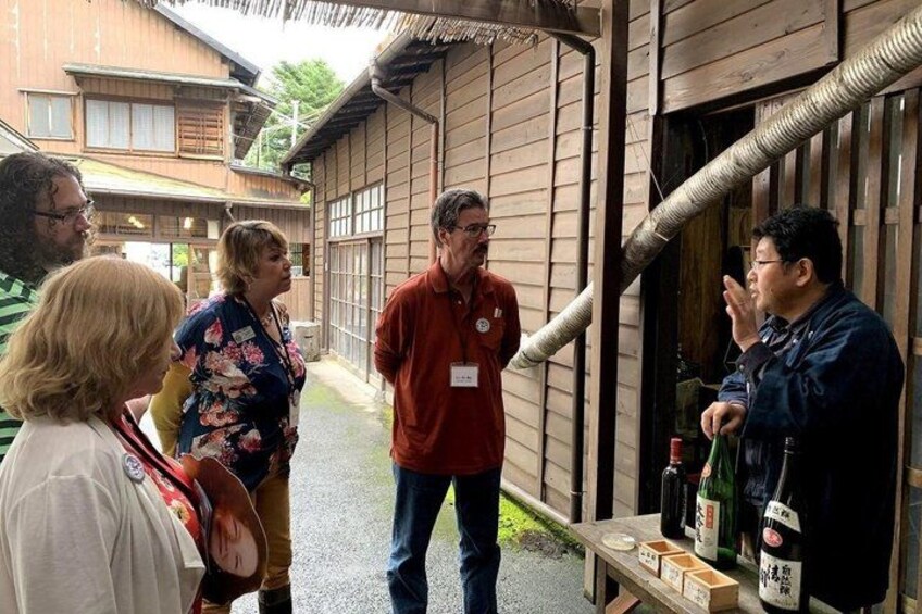 A group of people standing outside a wooden building with a metal pipe, a table with bottles, and a wooden bench. 5