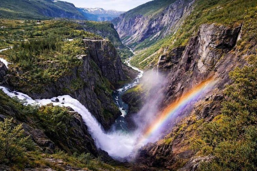 A waterfall with a rainbow, surrounded by green mountains and a river. 1
