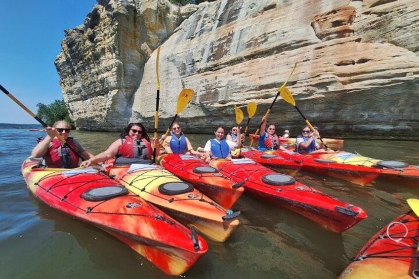 A group of people kayaking in red kayaks with paddles, near a rocky cliff. 2