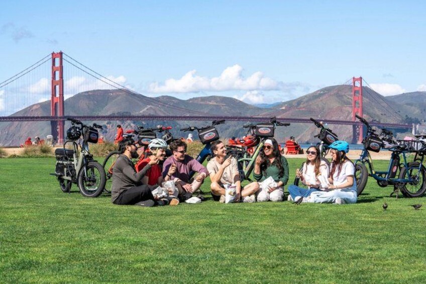 A group of people sitting on grass with bicycles and a bridge in the background. 2