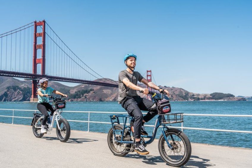 Two people riding bicycles on a bridge with a body of water and hills in the background. 3