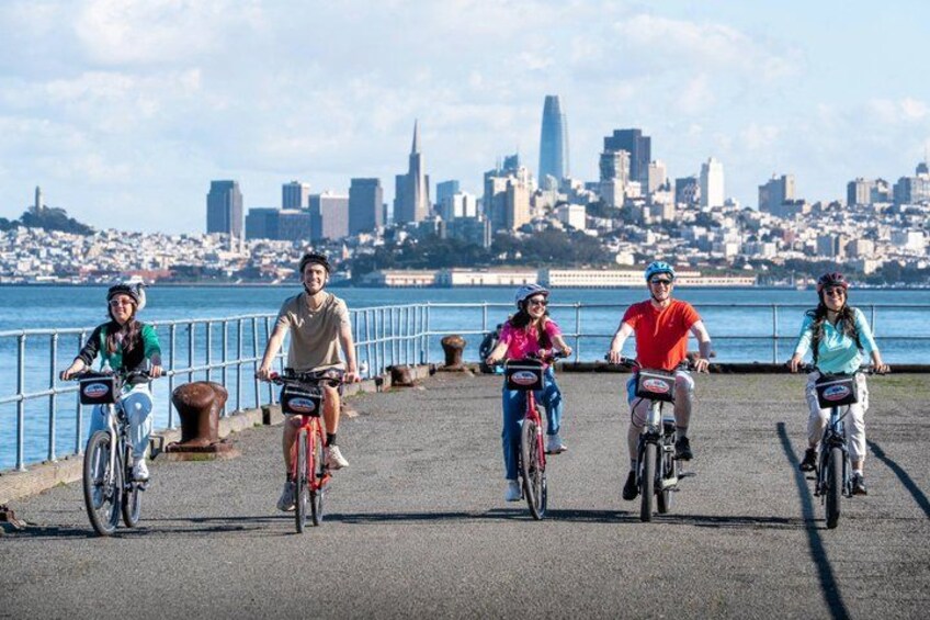 A group of people riding bicycles along a waterfront path with a city skyline in the background. 1