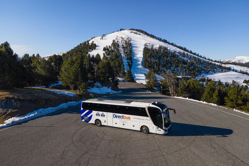 Un autobús blanco con la marca 'Directbus' circulando por una carretera con parches de nieve, rodeado de árboles y una montaña nevada. 2