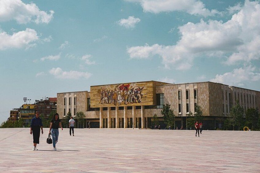 A large building with a mural, a paved plaza, and a few people walking. 4