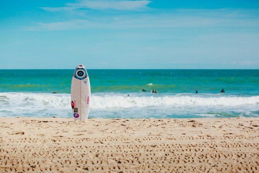 A beach with a surfboard standing upright in the sand, waves in the background, and a clear sky. 1