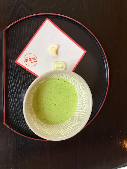 A green matcha bowl on a black tray with a red and white design, and a small piece of matcha on a white paper. 3