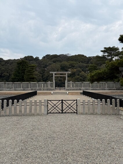 A traditional Japanese torii gate at the entrance of a shrine, surrounded by gravel and a fence, with a forested hill in the background. 4