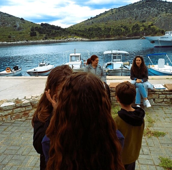 A group of people sitting on a stone wall by a lake with boats docked in the background. 1