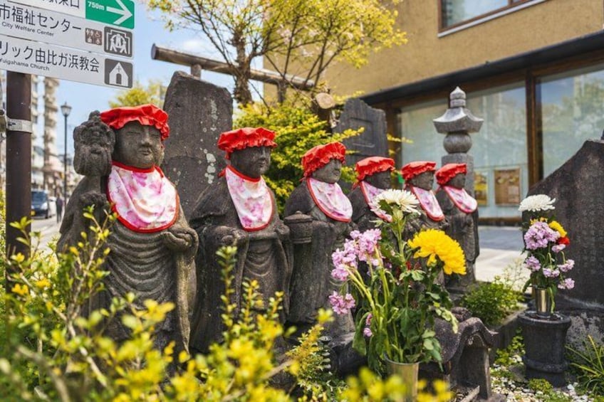 Stone statues with red hats and pink scarves, flowers, and a sign with Japanese text. 2