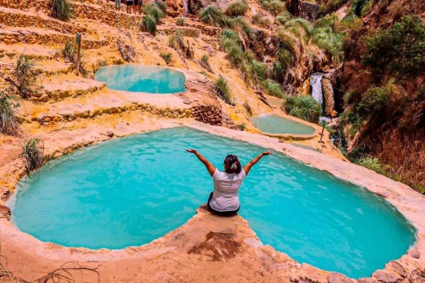 A person sitting in a turquoise pool with arms raised, surrounded by terraced fields and a waterfall in the background. 2