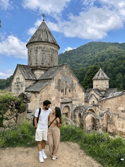 A historic stone church with a bell tower and a forested hill in the background. 1