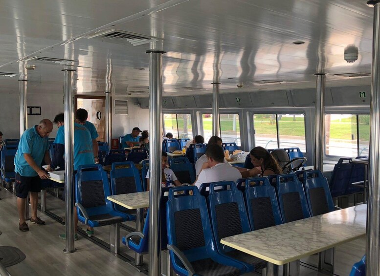 Interior de un ferry con sillas y mesas azules, pasajeros sentados y una persona sirviendo comida. 4