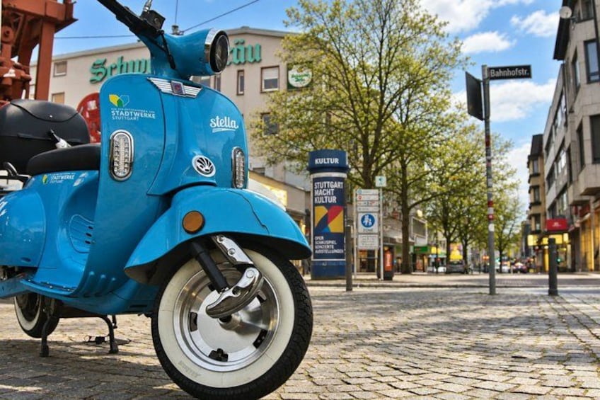 A blue scooter parked on a cobblestone street with buildings and a sign in the background. 4