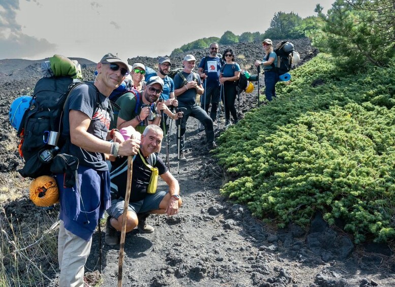 A group of hikers on a rocky trail with backpacks and hiking poles, surrounded by greenery and a hilly landscape. 1