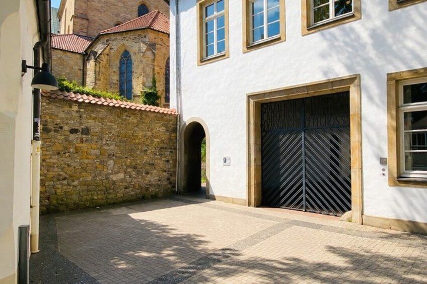 A paved courtyard with a stone wall, a large metal gate, and a building with arched windows. 3