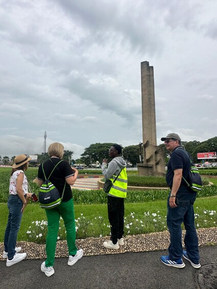 Eine Gruppe von Menschen steht in einem Park mit Grünflächen und bewölktem Himmel in der Nähe eines hohen Monuments. 4