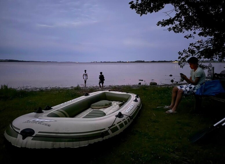 A riverbank with a green inflatable boat, a person sitting on a bench, and another person walking. 1