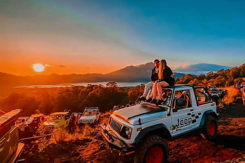 Valkoinen Jeep-merkkinen auto, jonka kyljessä on Jeepin logo, on pysäköity sorapintaiselle alueelle. Taustalla näkyy auringonlasku ja kauempana vuoria. 1