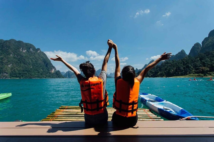 Two people on a boat wearing life jackets, with their arms raised, looking at a mountainous landscape and a calm sea. 1