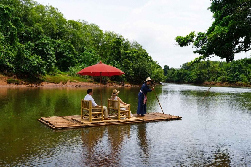 Un couple se trouve sur une plateforme flottante, abrité sous un parasol rouge, entouré d’une végétation luxuriante et bordé par une rivière paisible. 1