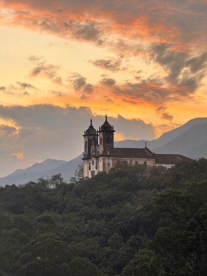 A church with two towers on a hill, surrounded by dense forest and mountains under a sunset sky. 1