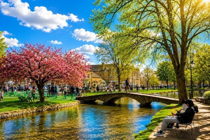 A park with a bridge over a calm river, cherry blossoms, and a person sitting on a bench. 5