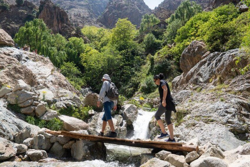 Two hikers crossing a narrow wooden bridge over a stream in a mountainous area with lush greenery. 4